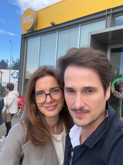 A man and a woman pose for a selfie. The man is on top with dark hair and a mustache, wearing a black collared jacket over a white shirt. The woman is below him, wearing glasses, a light-colored sweater, and a gray coat with a gold chain strap. Behind them is a building with a yellow overhang bearing the "SOL" logo. A sign with an image of a stylized fish is also visible. People walk past them, and there are red chairs and tables in the background. The location appears to be in Vagos, Portugal, based on the visible signage.