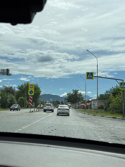 The image depicts a view from inside a vehicle traveling on a road in Ir, Russia. The road is paved and has multiple lanes. Several cars are visible on the road, including a white SUV, a white sedan, and a dark-colored sedan. Traffic signals and road signs are present, including pedestrian crossing signs and a "no entry" sign. Trees line the sides of the road, and mountains can be seen in the distance. The sky is cloudy with patches of blue. The overall impression is a typical urban or suburban street scene under a partly cloudy sky.