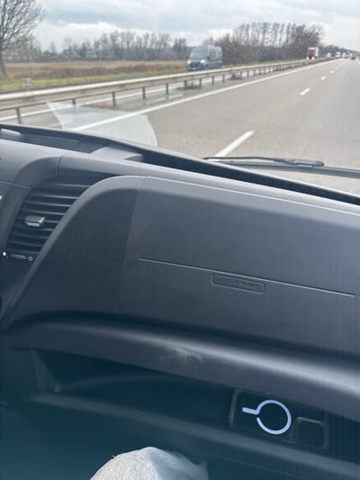 The image depicts a view from the interior of a vehicle, looking forward through a wet windshield onto a multi-lane highway near Niedernai, France. In the immediate foreground, the dark grey, textured dashboard of the vehicle is visible, featuring an air conditioning vent on the left and the embossed text "AIR BAG" on the passenger side. Below the main dashboard, an open storage cubby contains a transparent object with a prominent white circular design, possibly a mobile phone in a case with a MagSafe ring. A portion of a person's knee, clad in light grey fabric, is visible at the bottom left, indicating a passenger is present.

Beyond the windshield, the highway surface appears wet, reflecting an overcast sky with visible raindrops on the glass. A metal guardrail runs parallel to the road on the left, separating the highway from an adjacent field of brown, low-lying vegetation. A dark-colored van is parked or traveling slowly beyond the guardrail. In the far distance on the highway, multiple vehicles are visible, including a red truck. The horizon is lined with bare trees or dormant deciduous vegetation, suggesting a cool or winter season. The sky is uniformly grey and cloudy.