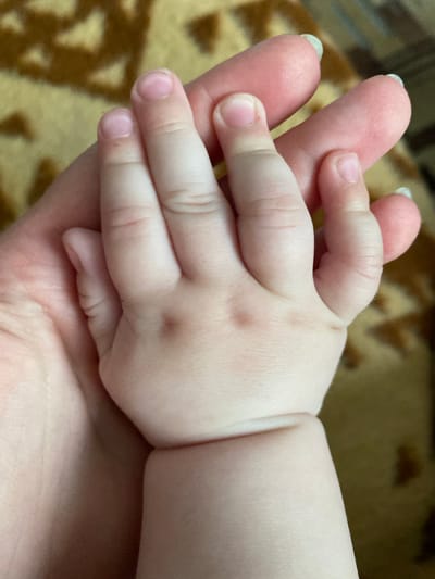 A close-up captures an adult hand holding a baby's hand. The adult's hand, with manicured nails, cradles the baby's hand, which has soft, plump skin. The baby's fingers are curled around the adult's fingers. The background is a blurred, patterned surface. The location is not discernible from the image.