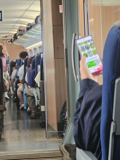 The image depicts the interior of a train carriage. In the foreground, a person in a dark blue jacket holds a smartphone displaying a grid of images, possibly a selection of plants or nature scenes, with Chinese text below. The phone screen is illuminated. To the left, other passengers are seated in blue train seats, some with luggage stowed above on overhead racks. The aisle is visible, with a checkered bag on the floor near one passenger. The train carriage appears to be in motion, as suggested by the blurred background and the overall lighting. The environment is utilitarian, designed for travel. The specific location in Yuhuatai District, China, is not explicitly evident from the visual cues within the image itself.