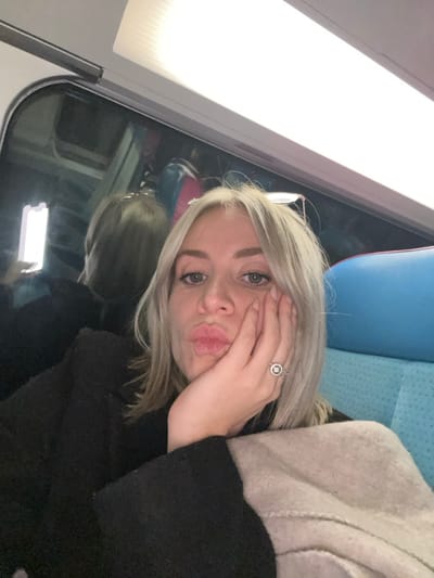 A woman with blonde hair is lying on a train seat. Her hair is blonde and slightly messy. She's wearing a black sweater and a beige jacket. She has her hand on her face, near her mouth, and is making a kissing face. She's wearing a silver ring with a large stone. The train window is behind her, with a blurry reflection of the interior. The seat appears to be blue. The overhead light is bright.