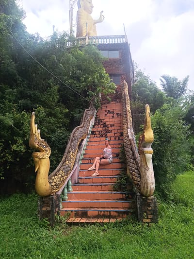 A woman is sitting on a set of ornately decorated, red-brick stairs leading up to a large golden Buddha statue in Thap Put, Thailand. The stairs are flanked by golden serpent-like structures that serve as handrails. Lush green vegetation surrounds the stairs. The sky is overcast.
