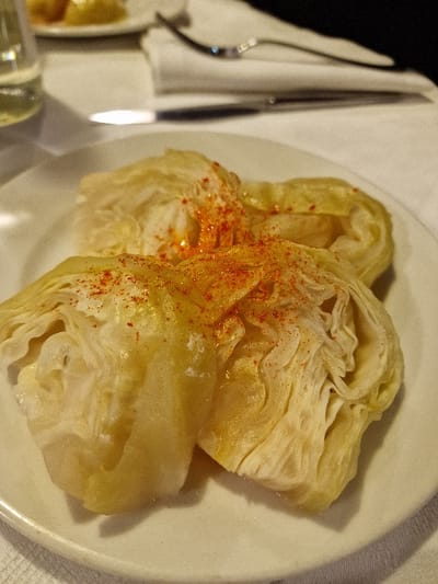 The image presents a close-up, elevated view of three portions of prepared cabbage, likely cabbage rolls or boiled cabbage leaves, arranged on a round, off-white ceramic plate. The cabbage portions are light yellow to off-white, exhibiting a slight sheen, and are generously sprinkled with a reddish-orange powdered spice, consistent with paprika. Visible folds and textures are present on the surface of the cabbage. The plate rests on a white, textured tablecloth. In the blurred background, indicative of an indoor dining environment, a partially visible clear glass bottle or carafe is present on the left. To the right, a folded white napkin holds a silver-colored fork and knife. A second, out-of-focus white plate containing yellowish food items is visible in the upper left background. This scene is located in a dining establishment within the city of Odorheiu Secuiesc, Romania. No individuals are visible.