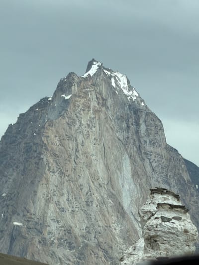The image shows a close-up of a jagged, grey mountain peak with patches of snow near its summit against a cloudy, overcast sky. In the foreground, a stack of white, weathered rocks, appearing almost like a natural sculpture, is partially visible. The larger mountain's face is textured with vertical striations and appears to be made of rock. There are no people or discernible human activity in the scene. The location is identified as Kurgiakh, India.
