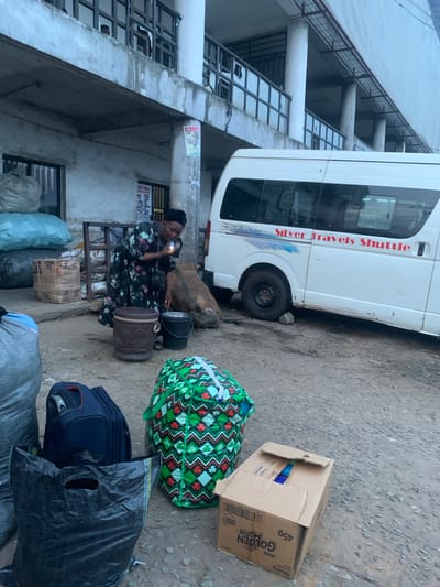 In the city of Elelenwo, Nigeria, a scene unfolds outside a building. A woman, dressed in a dark floral patterned dress and a headscarf, is bent over, holding a small object that appears to be a lighter near her face. She is standing on a dirt and gravel surface. In front of her are several objects: a dark brown, patterned bucket with a handle, and a black bucket. To the right of the buckets, a large sack made of rough material is partially visible. Behind the woman, a white shuttle bus with red lettering that reads "Silver Travels Shuttle" is parked. The bus has tinted windows and a silver stripe running along its side. Various bags and luggage are scattered around the scene. To the left of the woman, large, light blue and grey sacks are stacked. In the foreground, a navy blue suitcase and a large, green and white patterned bag are placed on the ground. A black plastic bag with handles is also visible in the foreground. Behind the shuttle bus, a large, rough sack is propped against it. A cardboard box with visible labels, including "GOLDEN MORN 45g", is also on the ground in the foreground. The environment appears to be an open area, possibly a bus station or market, with a building characterized by concrete columns and balconies in the background. The lighting suggests it is daytime, possibly overcast.