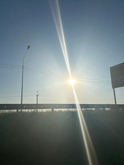 The image shows a bright sun shining in a pale blue sky, with rays extending outward. A signpost and utility poles are visible in the lower part of the image, along with a guardrail separating the road from a dark, overgrown area. The location is unspecified.
