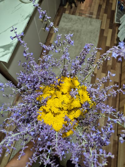 An overhead shot shows a bouquet of purple flowers with yellow flowers in the center. The bouquet is placed on a wooden floor. To the left of the bouquet, a white countertop with cabinets can be seen. In the background, there is a light gray rug and a dark wooden floor. The scene is indoors.
