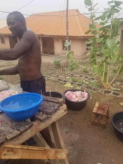 A man, shirtless and wearing black shorts, is preparing raw meat outside a house. He stands near a wooden table with a blue bowl of water and several pieces of raw meat on it. There are also two black plastic containers with raw meat. The man is in a dirt yard. There is a small wooden stool to the right and a single-story house in the background. There are multiple plants behind the house.  The location is in the city of Pati Ridge, Nigeria.