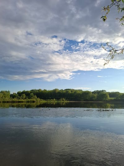 The image shows a view of a body of water, likely a lake or river, with trees lining the opposite bank. The sky is visible with clouds and blue patches. The image appears to be taken in Дерезовка, Russia. There are no people visible in the image. The scene appears to be calm and still.