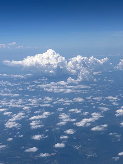The image is an aerial view of a landscape under a bright blue sky dotted with white, fluffy clouds. The clouds are varied in size and density, with a prominent cluster of larger clouds in the upper middle of the frame, and scattered smaller clouds across the lower half. Beneath the clouds, a dark blue and green landscape is visible, suggesting land with vegetation and possibly bodies of water. A winding road or river is visible cutting through the landscape, and some structures appear in the lower right portion of the image. The overall impression is one of a vast, serene aerial vista. The scene does not provide specific geographical markers to confirm the location in Viehhausen, Austria.