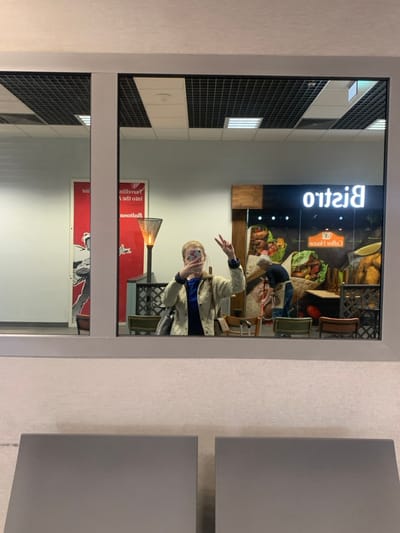 The image shows a reflection in a large glass window. The reflection is of the interior of a restaurant in Pyrzowice, Poland. The restaurant has a large sign above the counter, reading "Bistro", and a smaller sign that says "Coffee House". The restaurant is in a brightly lit area with a white ceiling with square ceiling tiles. There is a woman in the reflection, taking a selfie. She is wearing a beige jacket over a blue shirt. There is also a man reflected, standing behind a counter. He is wearing a grey shirt and blue jeans. There are several tables and chairs in the reflection. There is a large red advertisement poster on the left side of the reflection, and a white poster on the right side. The posters are both partially visible in the reflection. The image appears to be taken from the exterior of a building. Two grey chairs are in the foreground, partially blocking the bottom of the image. The image was captured at the coordinates 50.471° N, 19.073° E.
