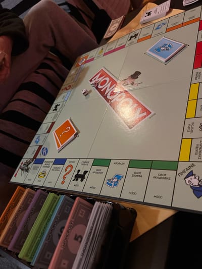 The image displays a Greek-edition Monopoly board game actively being played indoors within the Gerakas Municipal Unit, Greece. The light green game board is laid out on a flat surface, likely a table, under artificial lighting. Two white dice are resting near the center of the board, showing a roll of three and two dots. A black plastic tray in the foreground holds stacks of Monopoly game money in various denominations and colors, including pink (5), purple (20), green (100), and orange (50). Several game cards are present on the board: an orange stack identified as "ΑΠΟΦΑΣΗ" (Decision/Chance) and a blue stack identified as "ΕΝΤΟΛΗ" (Order/Community Chest). Property spaces feature Greek text, such as "ΟΔΟΣ ΗΡΟΔΟΤΟΥ" (Herodotus Street), "ΟΔΟΣ ΣΚΟΥΦΑ" (Skoufa Street), and "ΟΔΟΣ ΑΚΑΔΗΜΙΑΣ" (Akadimias Street). The lower body of an individual, wearing light grey sweatpants with distinct dark horizontal stripes, is visible to the left, indicating at least one participant. A partial view of another person's leg and arm is discernible in the upper left background, suggesting multiple players are present.