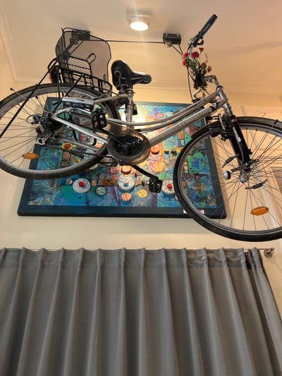 This image depicts an indoor scene in Saint-Gilles - Sint-Gillis, Belgium. A silver-colored bicycle, equipped with a black mesh front basket, a black rear rack, and black fenders, is suspended horizontally from a white ceiling via a black pulley system. Artificial red and pink flowers with green foliage are attached to the handlebars. A small, light-colored object is secured to the bicycle frame. Visible reflectors include an orange unit on the front wheel spokes and another on the rear. Directly beneath the bicycle, mounted on a light-colored wall, is a large, rectangular abstract artwork. The art features a dark frame and comprises a mosaic of blues, greens, yellows, and oranges, incorporating various circular and rectangular elements, some appearing three-dimensional. Below the artwork, grey, vertically pleated curtains are drawn, suspended from a visible curtain rod on the right. A round white ceiling light fixture is situated above the bicycle's mid-section. No individuals are present in the scene.