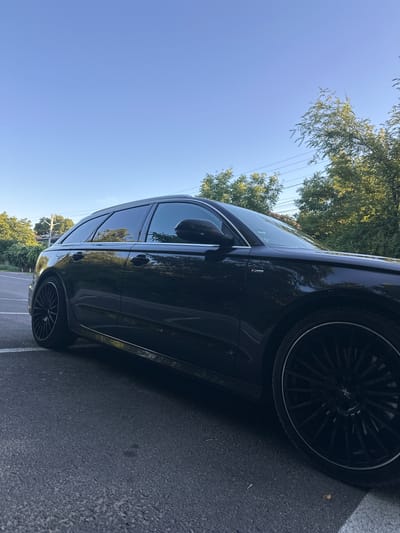 An Audi A6 Avant station wagon, finished in a dark color, is parked on an asphalt surface, likely a street or parking lot. The car is angled towards the viewer's left, with its front partially visible. The vehicle features tinted windows and custom black rims with a thin silver accent. The background consists of lush green trees and a clear blue sky with a few power lines visible. The scene appears to be in a suburban or semi-rural area, potentially on a sunny day. No people are visible in the image. The context of Orhei, Moldova is not discernible from the visual information presented in the image.