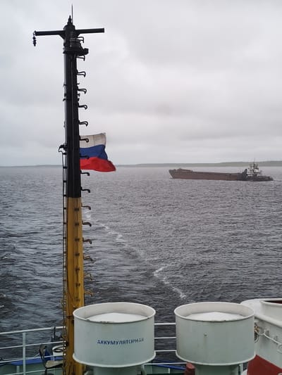 The image shows a view from a ship or boat. In the foreground are two cylindrical containers with the word "АККУМУЛЯТОРНАЯ" written on them. A metal pole with a ladder and a Russian flag attached to it is located to the left of the containers. In the background, there is a body of water with a ship or barge moving across it. The sky is overcast and grey. The location is likely near the city of Gorushka, Russia, given the Russian flag and Cyrillic text.