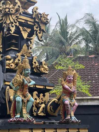 A tilted, ornate structure showcases Balinese art. Two horizontal figures, carved and brightly painted, adorn the black and gold framework. The top figure is pink, while the bottom is blue. Tropical palm trees and a tiled roof are visible in the background against a pale sky, indicating an outdoor setting.