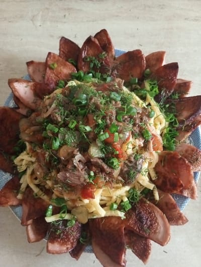 This media file is a top-down, close-up shot of a platter of food. The dish appears to be a hearty meal, possibly a traditional Russian dish, served on a blue and white patterned plate. The main components of the dish are noodles, shredded meat, and what looks like a stew or sauce with vegetables. The entire dish is garnished with chopped green onions and dill. Surrounding the central portion of the food are several slices of pan-fried sausage or meat, arranged in a circular pattern like petals. The meat slices are golden brown and glistening, indicating they have been cooked to a crisp. The lighting suggests this meal is either indoors or outdoors on a bright day, with no specific time of day or weather conditions discernible. The background is a light-colored, textured surface, likely a table, with a slightly blurred wooden grain pattern. There is no text visible in the image. The scene evokes a sense of a home-cooked meal, perhaps a family gathering or a special occasion. The focus is entirely on the appetizing presentation of the food.