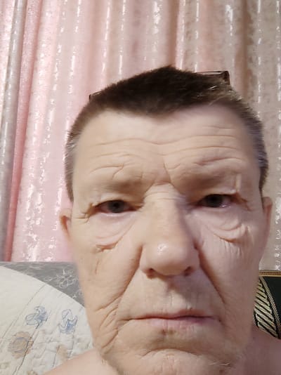 The image is a close-up, portrait-style photograph of an older individual's face. The subject has short, dark hair and is looking directly at the camera. Prominent wrinkles are visible on their forehead and around their eyes and mouth. The background consists of pink, textured curtains and a portion of a patterned duvet. The lighting is overhead, creating shadows that emphasize the facial features. The specific location within Бәйтерек, Kazakhstan, is not discernible from the image itself.
