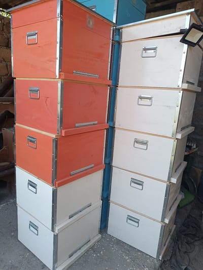 In Ghukasavan, Armenia, a vertical, eye-level medium shot captures a stack of wooden boxes with metal reinforcements and handles. The boxes are painted in alternating shades of orange and white, with some in light blue visible in the background. The scene is outdoors, with a rough concrete ground surface and a backdrop of stacked logs and a stone wall. A black LED floodlight is attached to one of the boxes on the right. The overall impression is one of organized storage or a workshop setting.