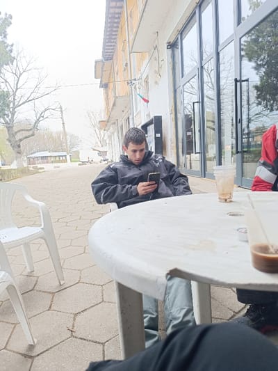The image depicts an outdoor seating area in Vinitsa, Bulgaria, under an overcast sky. A young male with short dark hair, wearing a dark jacket and light-colored pants, occupies a white plastic chair at a round white plastic table in the midground. He is focused on a black smartphone held in both hands. To his right, an individual in a red and black jacket is partially visible, also seated at the table. An empty white plastic chair is positioned to the left. On the table, a clear plastic cup with light brown liquid residue and a partially visible plastic cup containing a dark brown liquid with a white creamer packet are present. The ground is paved with hexagonal concrete tiles. In the background, a two-story building with a faded light-colored facade, featuring multiple windows and a small Bulgarian flag, stands. Beyond this, bare trees, a utility pole, a paved path, and a white van are visible in the distance. The lower legs of a third individual, clothed in dark fabric, are in the extreme foreground.