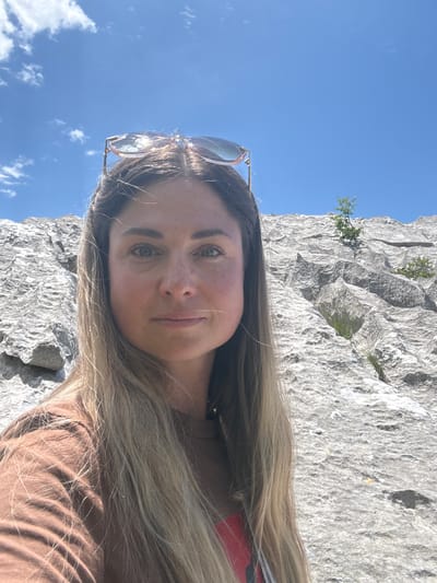 A woman is taking a selfie outdoors with a rocky landscape and a bright blue sky in the background. She has long blonde hair and is wearing a brown t-shirt and sunglasses perched on her head. The rocky terrain appears to be a natural formation, possibly a cliff face, with sparse vegetation growing in some crevices. The lighting suggests it is a sunny day. The location is the city of Žlijebi, Montenegro.