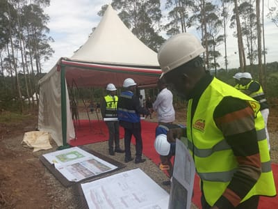 This image captures an outdoor gathering at what appears to be a construction or development site in Moticho ward, Kenya, during the daytime under a partly cloudy sky.
In the foreground, several men are present, most wearing white hard hats and high-visibility safety vests, indicating roles in construction, engineering, or project management. The most prominent figure, in the right foreground, wears a cream-colored hard hat and a neon yellow safety vest over a brown and black striped long-sleeved shirt. He is intently examining large-format documents, likely architectural or engineering plans, spread out on the ground. Other plans are also visible on the ground nearby.
Behind him, another man in a white hard hat and a blue jacket with "YOOLIANCES LTD" (partially visible) on the back is also looking at the plans. Further in the background, several other individuals are gathered near a large white canvas tent with a pointed roof. The tent is adorned with red, white, and green fabric elements, possibly representing the Kenyan national colors, and a red carpet leads into its entrance.
The ground is a mix of reddish-brown soil and gravel. Tall, slender trees, possibly eucalyptus, densely populate the background, suggesting a semi-rural or forested setting. The overall scene suggests a site visit, project briefing, or a formal groundbreaking ceremony for a construction project.
Visible text includes "GISAMO HOLDINGS LTD" on the back of the foreground man's safety vest and various diagrams, layouts, and text on the architectural plans, though most of the text on the plans is not clearly legible.