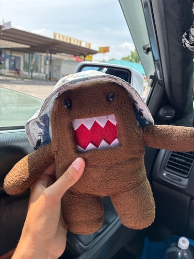 A close-up shot captures a person's hand holding a brown Domo-kun plush toy inside a vehicle. The toy wears a bucket hat adorned with a dinosaur pattern. The background reveals a street scene with buildings and a bright sky, suggesting an outdoor location in Chernigovka, Russia. The toy is positioned in the foreground, partially obscuring the car's interior, including the dashboard and air vent. The lighting indicates a sunny day.
