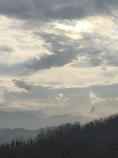 An outdoor landscape shot, presumably taken from Villa di Gemmano, Italy, depicts a panoramic view dominated by a complex sky and layered terrain. The upper two-thirds of the image are filled with a heavily overcast sky, composed of numerous gray and white stratocumulus clouds. A prominent, bright white aperture in the cloud cover is visible in the top-center to top-right quadrant, indicating a direct sunlight source. Below the sky, the landscape features multiple successive ridges of hills or mountains, receding into the distance and exhibiting atmospheric haze that renders them in progressively lighter shades of blue-gray. The immediate foreground, occupying the lower third of the image, is characterized by a dark, densely forested hillside. The trees appear to be predominantly deciduous, standing bare, suggesting a dormant season. No human figures, animals, or distinct man-made structures are identifiable within the frame.