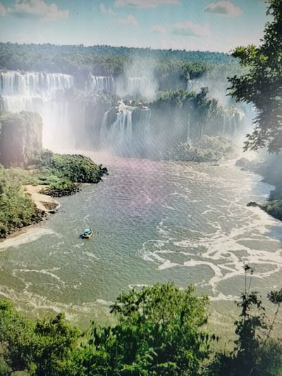 This is a high-angle, full shot of the Iguazu Falls, a natural wonder located on the border of Argentina and Brazil. The image captures a wide expanse of cascading waterfalls amidst lush, green, forested cliffs. A wide, calm river flows in the foreground, with a small boat visible on the water. The sky above is blue with scattered white clouds. The scene is a majestic display of natural beauty, with the mist from the falls creating a soft, ethereal atmosphere. There is no visible connection to Yashkino, Russia in this image.
