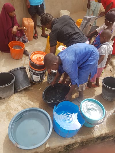 The image shows a group of people in Dutsinma A, Nigeria, collecting water. Several individuals are positioned around a water source, filling various buckets and containers. A child in a blue outfit is actively filling a black bucket. Other containers nearby include black, blue, and orange buckets. Some are filled with water. One woman is sitting beside a container. The ground around the water source appears wet, indicating recent spillage or ongoing activity.