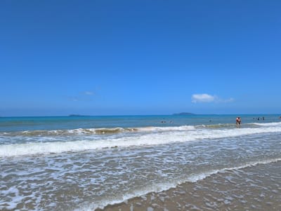 The image depicts a wide, sandy beach with gentle waves rolling onto the shore under a clear, bright blue sky. In the distance, the turquoise ocean stretches to the horizon, meeting a hazy sky. A few small islands or landmasses are visible on the far horizon. Several people are in the water, some swimming and others standing. One person, a man, is standing in the water near the shore, facing the camera. The overall scene is one of a sunny day at the beach, with tranquil waters and a sense of peaceful recreation. The provided context states the location is Tianya District, China, which is a district in Sanya, Hainan Province.