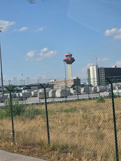 The image shows an airport control tower and several buildings under a partly cloudy blue sky. In the foreground, a chain-link fence runs across the frame, with dry grass and weeds behind it. Beyond the fence, a row of white, covered cargo containers on trailers is visible, possibly at a cargo terminal. In the distance, the distinctive control tower with a red top stands tall, flanked by several other airport buildings. The scene appears to be an outdoor, daytime environment, likely at an airport. The location is identified as Walldorf, Germany.
