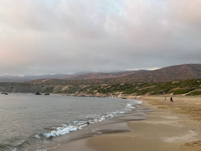 This is a photograph of a cloudy day at a beach in Ineia, Cyprus. The scene is dominated by a calm, gray ocean with gentle waves lapping onto a sandy shore. In the distance, a hilly landscape covered in dry brush and some greenery stretches across the horizon, with a few scattered structures visible on the highest points. The sky is overcast with thick, gray clouds, suggesting either early morning or late afternoon, or possibly impending rain.

Several people are visible on the beach, appearing to be enjoying a leisurely day. Some are walking along the shoreline, while others seem to be standing or observing the water. The atmosphere appears relaxed and tranquil, with no indication of specific events. A small, rustic wooden shelter is situated on the edge of the beach, near the vegetation. The overall impression is one of a peaceful, natural coastal environment under a muted sky.