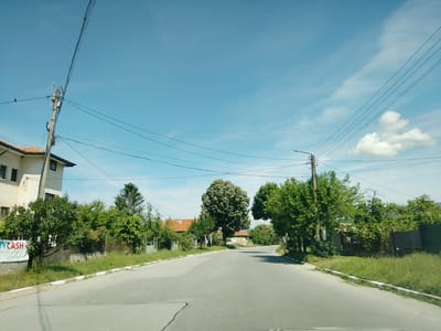 The image depicts a street scene in the city of Mirovo, Bulgaria, on a clear day with a bright blue sky and scattered clouds. The perspective is from a vehicle traveling along a paved road. On the left side of the road, a multi-story building with white walls and dark-framed windows is visible. Adjacent to the building is a banner with the text "CITYCASH". Further along the left side are several trees and a fence.

The road itself is gray asphalt with white and gray painted curbs. Overhead, a complex network of electrical wires and telephone lines crisscrosses the sky, supported by wooden utility poles.

On the right side of the road, there is a continuous line of lush green trees and vegetation. In the distance, more houses with red tile roofs can be seen amongst the trees. A white van with some visible markings is parked on the right side of the road in the mid-ground. The overall impression is a suburban or semi-rural setting.