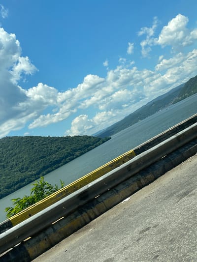 An outdoor scene, photographed from a tilted perspective, captures a roadside view overlooking a large river with forested landscapes and a partially cloudy sky. In the foreground, a segment of gray asphalt road, exhibiting minor wear and dark stains, occupies the lower right section. A two-tiered gray metal guardrail, showing evidence of weathering and grime, extends diagonally across the lower half of the frame. This guardrail is mounted on a barrier with extensively chipped and faded yellow paint, revealing underlying material and dirt, along with residual black and yellow striped markings at its base. A cluster of vibrant green leafy vegetation is visible emerging from behind the guardrail near the lower left.
Beyond the guardrail, a broad expanse of dark blue-green water, identifiable as the Danube River, stretches horizontally. On the left side of the river, a densely forested hill or mountain rises prominently from the water's edge. Along the right bank, a series of receding, tree-covered hills extends into the distance, with small clusters of buildings or settlements discernible along the waterfront. The sky above is a bright blue, featuring numerous white cumulus clouds scattered throughout. The lighting indicates clear daytime conditions. No individuals, specific vehicles, or direct human actions are depicted. The scene presents a combination of natural geography and roadside infrastructure, consistent with the environment in and around Drobeta-Turnu Severin, Romania.