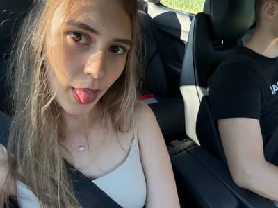 The image shows two people in a car, with the camera angled downwards. The person in the foreground, on the right, is a woman with long blonde hair. She is wearing a white top and has her tongue sticking out. She is also wearing a necklace with a small diamond pendant. The person in the background, on the left, is a man wearing a black t-shirt. He appears to be the driver of the car. The car's interior is visible, including the black leather seats and a seatbelt. The scene appears to be set during the day, with sunlight coming through the windows. The background outside the car shows greenery, suggesting a rural or suburban location. The overall mood of the image is playful and casual. The visible text on the man's t-shirt is "IA". The location context is Iffezheim, Germany.