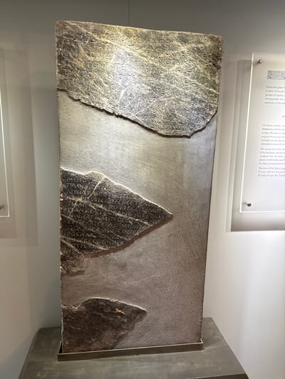 The image shows a large, rectangular stone stele with inscriptions. The stele is dark gray and appears to be made of marble or a similar stone. It is displayed vertically on a stand in what appears to be a museum setting. There are two main sections of inscriptions visible, with a large piece missing from the upper right and a smaller piece missing from the lower right. The text is carved into the stone in an ancient script.

To the right of the stele, a small information panel is visible, made of clear acrylic with text printed on it. The panel has a title that reads "Hymn". The text on the panel describes the stele as an "Inscribed marble stele from Palaikastro with the Hymn to Zeus". It mentions that the hymn was sung by young men, included circular dances, and was intended to ensure fertility. It also notes that the young men invite Zeus and his followers to join them, and the hymn is connected to events from Zeus's birth and subsequent life. The panel also mentions the form of the letters suggesting a date of "4th-early 3rd cent. BC".

While the city of Heraklion, Greece, is mentioned in the prompt as a potential location, this information cannot be visually confirmed from the image itself. The environment appears to be an indoor exhibition space with neutral-colored walls and subtle lighting. No people are visible in the image.