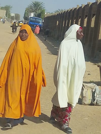 This image depicts an unpaved street scene in Mariri, Nigeria, under bright daylight. In the foreground, two individuals, presumably adult females, walk towards the viewer. On the left, a person wearing a voluminous, bright orange full-body covering (jilbab or abaya) with a hood covering the head, looks directly forward. This individual wears dark closed-toe shoes with white soles. To the right, another person wears a white full-body garment with a hood, revealing a red and white striped head covering underneath. This person's face is in profile, looking towards the right, and they wear patterned red and white pants under the white garment, with grey open-toed sandals.
The ground is dusty and unpaved, with long shadows cast towards the viewer, suggesting strong sunlight. In the midground, several vehicles are visible: a blue truck parked or moving on the left side of the street, a yellow tricycle (keke Napep), and a person on a motorcycle. Other indistinct figures are present in the distance. Along the right side of the street, a light brown concrete wall with repeating vertical indentations extends into the background. A large grey bag, possibly containing cement or grain, rests on the ground near the base of this wall. Palm trees and other buildings are visible in the far background.