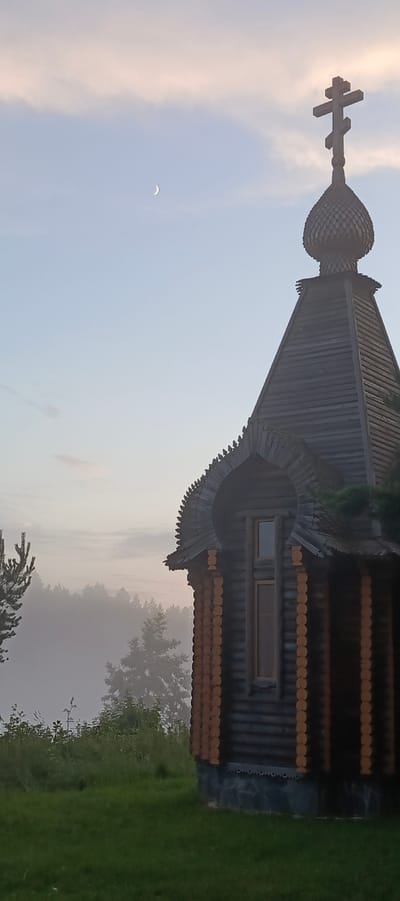 The image depicts a small, wooden Orthodox chapel situated in the city of Merchalovo, Russia. The chapel has a dark wood exterior, characterized by horizontal logs and decorative wooden trim along the roofline. A prominent feature is the onion-shaped dome topped with a multi-barred cross. The chapel is nestled amidst a grassy clearing, with a dense forest shrouded in mist visible in the background. The sky is pale blue with wispy clouds, and a sliver of a crescent moon is visible in the upper left quadrant. The overall atmosphere is serene and tranquil, suggesting an early morning or late evening setting due to the soft lighting and the presence of mist. No people are visible in the scene.