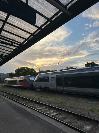 This is a photograph taken at the train station in Alès, France, likely in the late afternoon or early evening, judging by the lighting. The scene captures two passenger trains parked on parallel tracks. The train on the left is red and silver with yellow and orange accents, while the train on the right is predominantly silver with blue trim. Both trains appear modern and are empty. The viewpoint is from a platform, suggesting the observer is a passenger or staff member. Above the trains, the sky is visible with a mix of blue sky and scattered clouds, illuminated by the setting sun which casts a warm glow. A portion of the station's covered platform, constructed with a translucent, textured roof supported by dark metal beams, occupies the upper left quadrant of the image. The tracks and the gravelly area between them are clearly visible. The environment appears to be an outdoor railway station with some vegetation and buildings visible in the background.