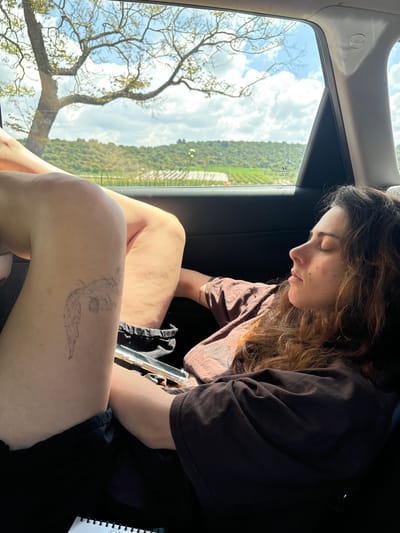 A young woman is seated in a vehicle, possibly a car, with her head tilted downwards, and her eyes closed, as if she is sleeping or resting. She has long, wavy brown hair and is wearing a brown short-sleeved shirt. Her legs are visible, with a tattoo of a butterfly or winged creature on her right knee. The vehicle is moving and the window shows a landscape with trees, bushes, and grass. The vehicle may be in Alona Regional Council, Israel.