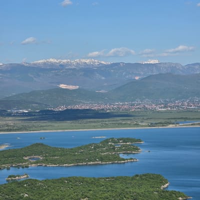 The image depicts an aerial view of the region surrounding the city of Ponikvica, Montenegro, with a focus on the Lake Skadar area.
To the left, a mountain range with some visible snow caps forms the backdrop. A town or city is nestled between the mountains and a green plain, possibly representing Ponikvica. This plain is bordered by a body of water, likely a section of Lake Skadar, which extends to the right side of the image.
Within the lake, several forested islands are visible, adding to the complexity of the landscape. The general environment appears to be a mix of mountainous terrain, urban or semi-urban development, and natural, vegetated areas surrounding a lake.