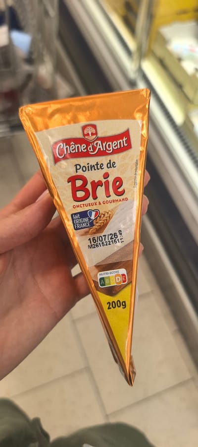 A wedge-shaped piece of Brie cheese, wrapped in orange foil, is held in a person's hand. The foil packaging features branding for "Chêne d'Argent Pointe de Brie," indicating it is "ONCTUEUX & GOURMAND" (creamy and gourmet). Below this, a small French flag symbol is present with the text "lait ORIGINE FRANCE" (milk origin France). A date "16/07/26" and a batch code are visible. A Nutri-Score is displayed, showing a rating of "D." The weight of the product is indicated as "200g." The background appears to be a retail environment, possibly a grocery store aisle, with shelves and other products blurred in the distance. The lighting suggests indoor illumination. The location is not specified in the image itself; however, if the context of Villeneuve-le-Roi, France, is assumed, this would be a common scene in a supermarket within that city.