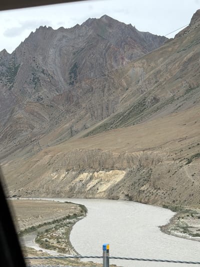 A wide, muddy river flows through a valley with steep, rocky mountains on either side. The mountains are layered with different shades of brown and gray rock, showing signs of erosion and geological folding. Sparse vegetation, appearing as small green shrubs and patches of dry grass, clings to the slopes. In the foreground, a portion of a dark frame, possibly from a vehicle window, obscures the lower left corner of the image. Several thin, parallel wires stretch across the lower part of the frame, with a small blue and yellow marker visible on one of them. The sky is overcast and a pale gray. The scene depicts a natural landscape with no visible human activity or structures, suggesting a remote, mountainous location such as the city of Pidmo, India, known for its dramatic terrain and river valleys.
