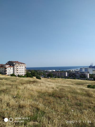 This is an exterior, daytime, eye-level, medium shot photograph taken from a slightly elevated position overlooking a coastal city. The foreground is dominated by a dry, grassy hillside covered in sparse, golden-brown vegetation. Several small pieces of litter are scattered across the hillside. In the mid-ground, a cluster of multi-story apartment buildings is visible on the left, with a large, cream-colored building featuring multiple balconies and a reddish-brown roof. To the right of the apartment buildings, a section of the city unfolds with more buildings, including some industrial structures with white cylindrical tanks and tall blue cranes that suggest a port or industrial zone. The background features a wide expanse of calm, blue water, likely the sea, with a clear, light blue sky above. The horizon line is clearly defined between the water and the sky. The scene appears to be located in Avcılar, Turkey, indicated by the text overlay. The overall impression is a partly developed urban landscape adjacent to a body of water.