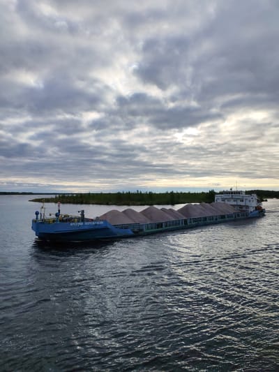A barge, loaded with gravel and emblazoned with Cyrillic script reading "АРСЕНИЙ ВОРОЖЕЙКИН" ("ARSENIY VOROZHEIKIN"), is moving across a body of water. The barge's cargo is arranged in multiple uniform mounds. In the background, a shoreline is visible with dense green vegetation. The sky is overcast with layered, gray clouds, suggesting a cloudy day or approaching dusk. The water shows gentle ripples, indicating a slight breeze. The scene appears to be a waterway possibly near the city of Bereg, Russia, given the Cyrillic inscription on the barge.