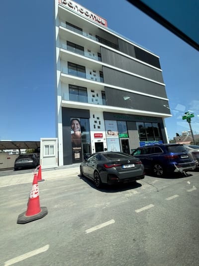 The image shows a modern, multi-story building with the name "pandemus" visible in red lettering at the top. The building has a white facade with dark, slatted panels on the upper floors and balconies with glass railings. Below, the ground floor features storefronts with large windows. One storefront displays a large advertisement with a woman's face and the text "Wellnest." Another storefront has signage that includes Greek and English text, reading "ΦΑΡΜΑΚΕΙΟ ΑΠТЕКА PHARMACY" and a green "OPEN" sign. A blue SUV and a dark grey sedan are parked in front of the building. In the foreground, two orange traffic cones are placed on the asphalt. The scene appears to be in a commercial area with a clear blue sky overhead. The location is identified as the Mouttagiaka Community, Cyprus, based on external information.