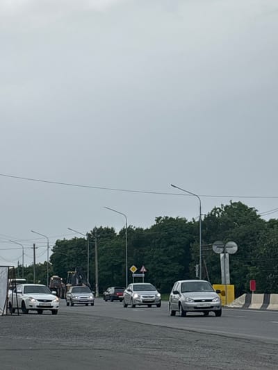 The image depicts a traffic scene on a cloudy day in Gizel, Russia. A line of white vehicles, appearing to be vans or minivans, are driving along a wet asphalt road, suggesting recent rain or ongoing dampness. The vehicles are moving in the same direction, creating a sense of a procession or convoy. The surrounding area is characterized by lush green trees that form a dense backdrop on the left side of the road. Several traffic lights and streetlights are visible, indicating an urban or suburban setting. Road signs, including a yellow diamond-shaped sign with a black symbol and a red rectangular sign, are present.

One of the vans has a license plate with Cyrillic text, suggesting it is Russian. The sky is overcast, casting a diffused light over the scene, and the overall mood is neutral, typical of a routine traffic situation. The focus is on the vehicles and the road, with the natural elements of trees and sky providing context. There is no clear indication of any specific event or activity beyond normal traffic flow. The perspective is from a slightly elevated position, looking down the road at the line of vehicles.