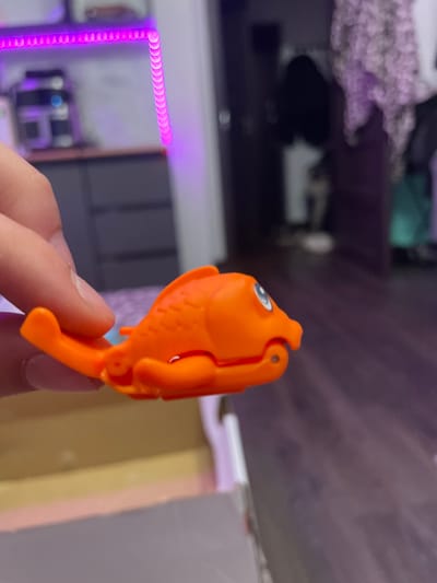A hand is holding a small, orange toy fish. The toy fish has a blue accessory attached to its underside. The background is blurred and appears to be an indoor environment with purple LED strip lighting visible in the upper left corner. There are cabinets and what appears to be a doorway in the background. The image is taken from a close-up perspective, focusing on the toy fish and the hand holding it.